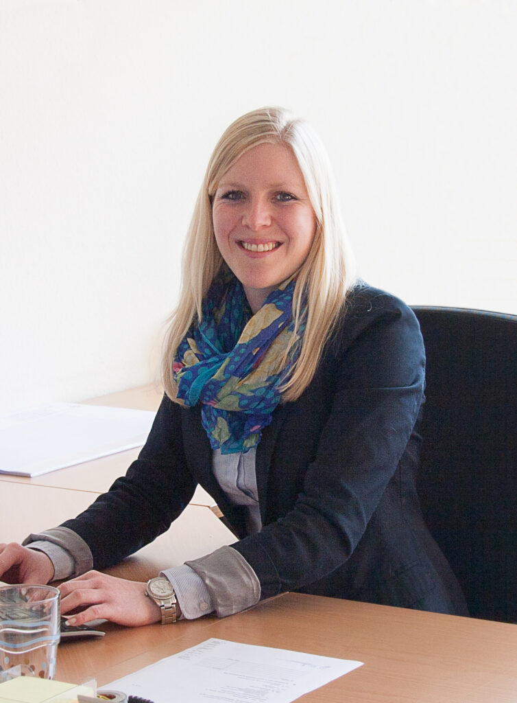 Foto von einer Frau mit langen, blonden Haaren am Schreibtisch. Sie trägt Blazer, gemusterten Schal und Uhr. Vor ihr liegen Papiere und ein Glas Wasser.