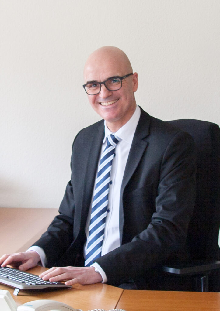 Foto von einem Mann mit Anzug, gestreifter Krawatte und Brille, der an einem Schreibtisch im Büro auf eine Tastatur tippt.