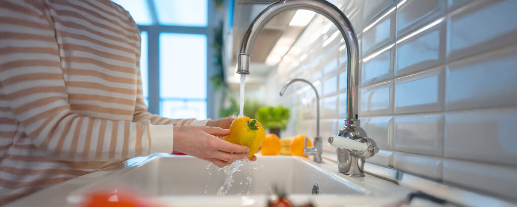 Foto von einer Person im gestreiften Shirt, die eine gelbe Paprika unter Wasser am Spülbecken wäscht; Obst und Gemüse im Hintergrund.