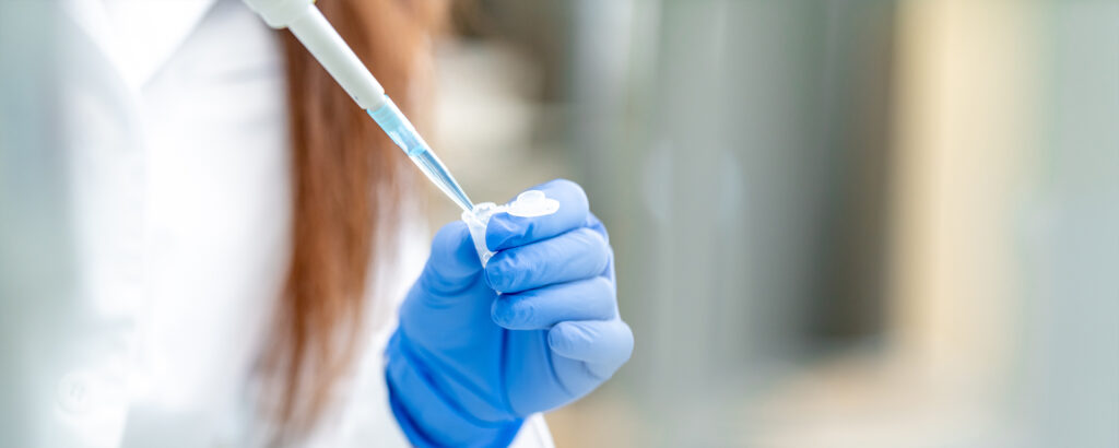 Foto von einer Person mit blauen Handschuhen, die mit einer Pipette Flüssigkeit in ein kleines Laborglas füllt.
