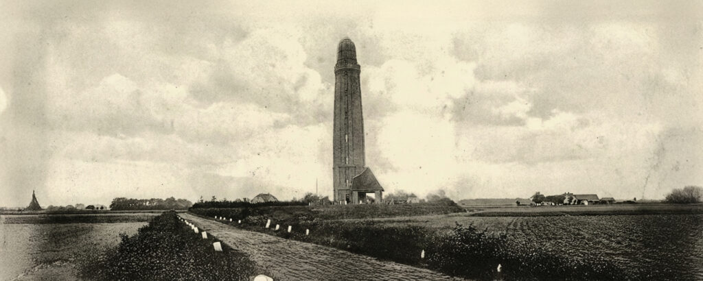 Schwarz-weiß-Foto von einem hohen Leuchtturm auf flacher Landschaft, Straße führt dorthin, Felder und Gebäude im Hintergrund, bewölkter Himmel.