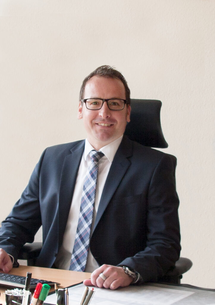 Foto von einem Mann mit Anzug und Brille an einem Schreibtisch im Büro. Auf dem Tisch liegen Stifte und Büromaterial. Heller Hintergrund.